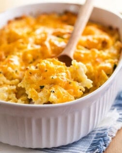 A white casserole dish with a wooden spoon scooping up Cracker Barrel hash brown casserole topped with cheese.