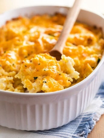 A white casserole dish with a wooden spoon scooping up Cracker Barrel hash brown casserole topped with cheese.