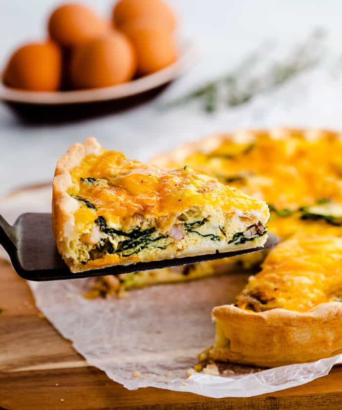 a pie cutting holding up a slice of ham and cheese quiche with a bowl of eggs in the background.