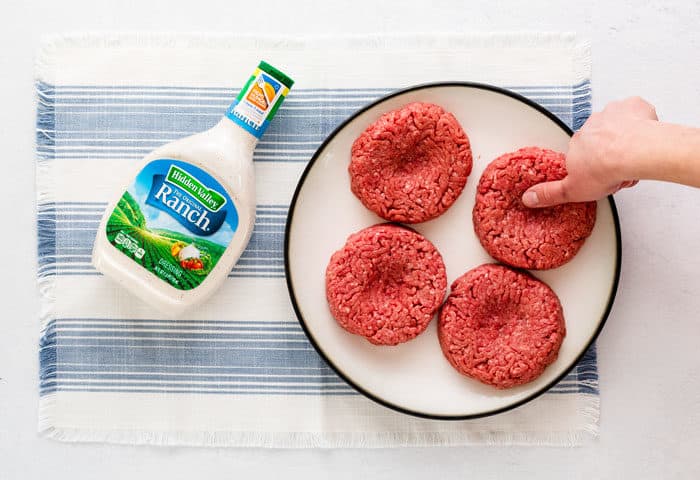 A plate with 4 raw burger patties next to a bottle of ranch salad dressing. A thumb is being pressed to indent the middle of the burgers.