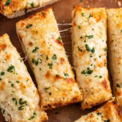 Sticks of cheesy garlic bread topped with parsley on a wooden cutting board.