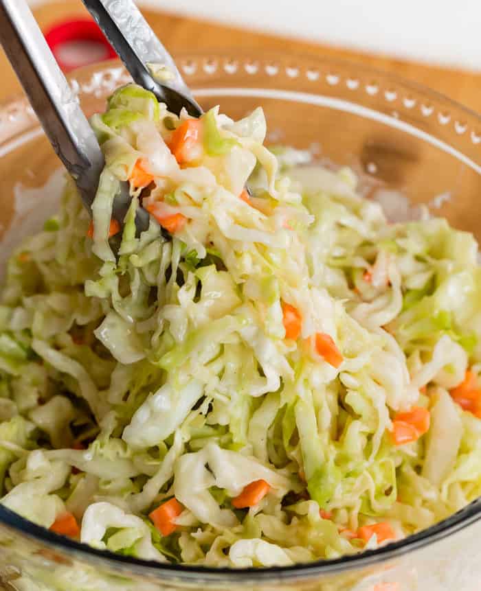 Metal Tongs Pulling up Copycat KFC Coleslaw from a glass bowl.