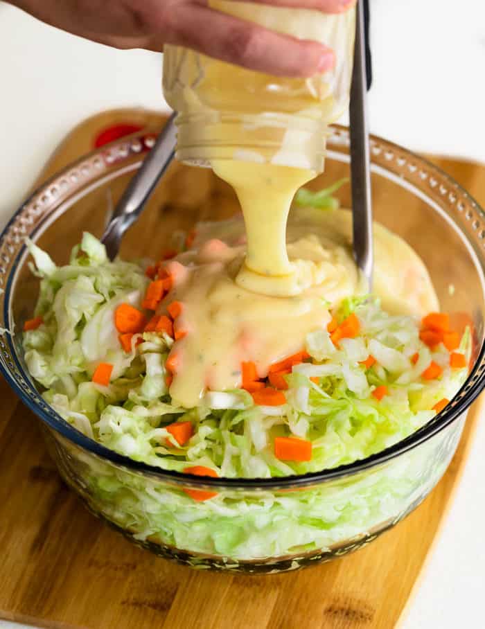 A hand holding a glass jar over a glass bowl of Copycat KFC Coleslaw and pouring the KFC Coleslaw sauce on top.