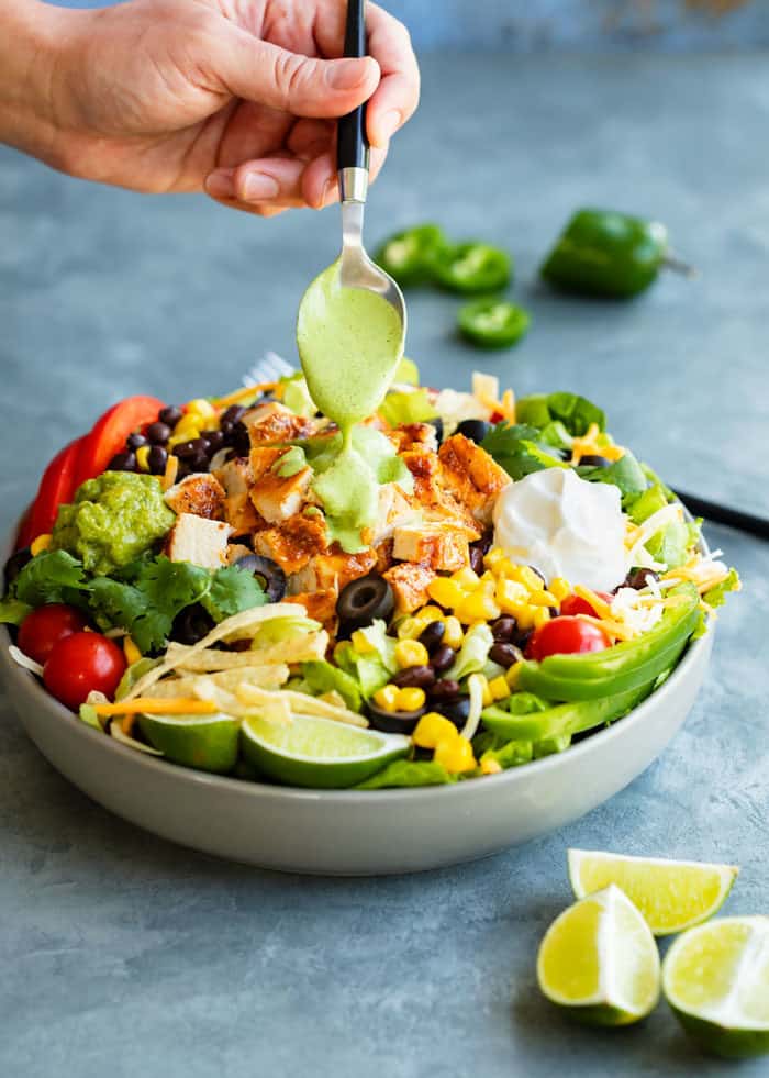 A hand holding a spoon and drizzling cilantro lime dressing over a big bowl of salad.