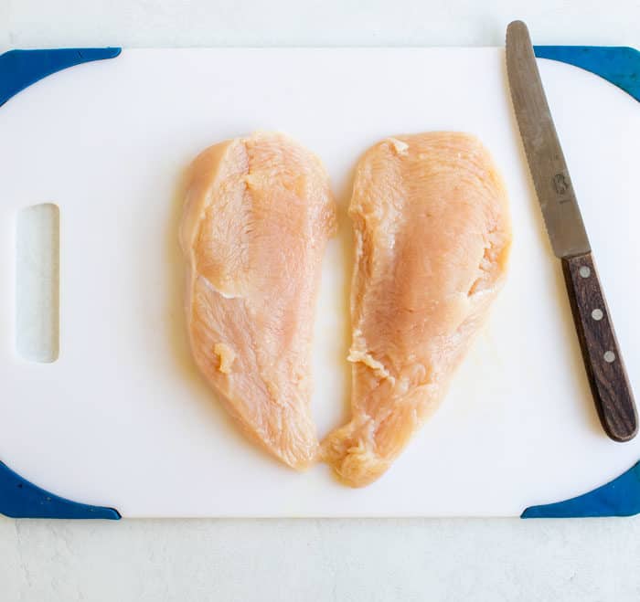 A halved Chicken Breast on a white cutting board next to a knife.