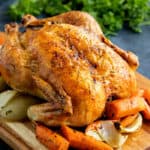 A roast chicken on a board with carrots and onions.