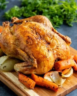 A roast chicken on a board with carrots and onions.