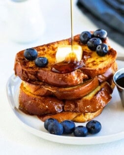 Syrup being poured over a stack of Alton Brown's french toast with blueberries.