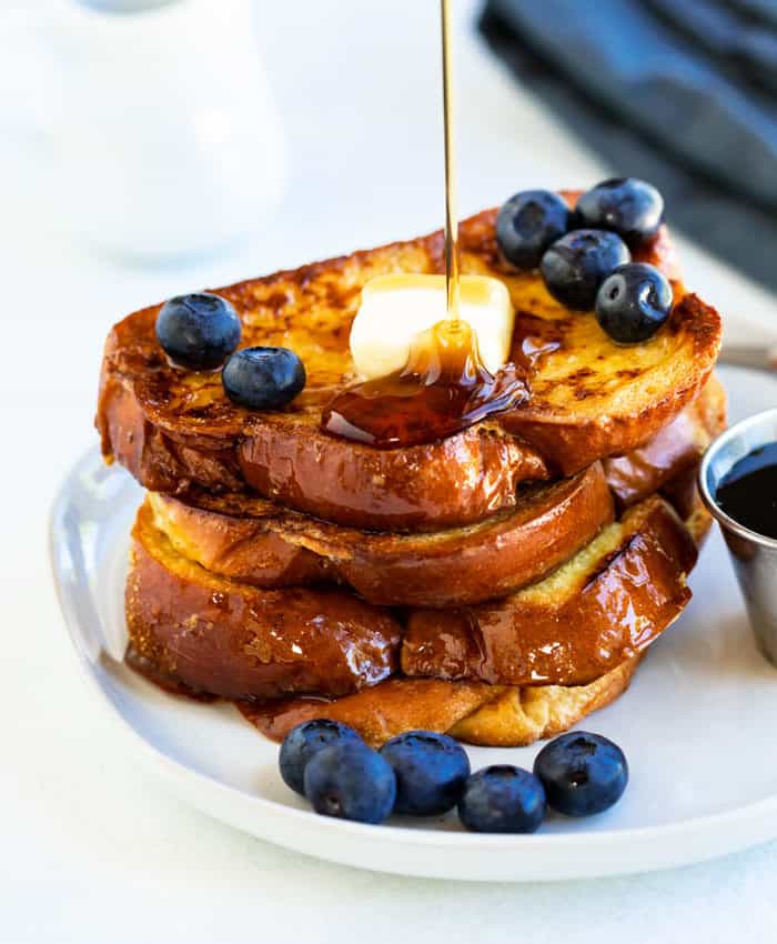 Syrup being drizzled over a stack of French Toast with blueberries and butter on a white plate.