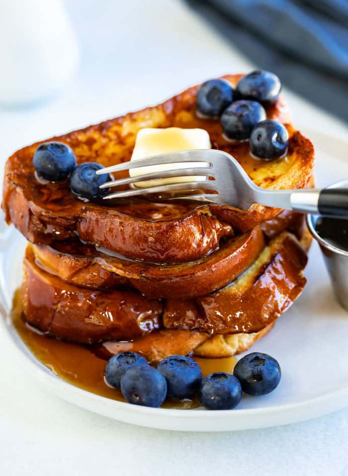 A fork cutting into a stack of french toast with blueberries and butter on a white plate.