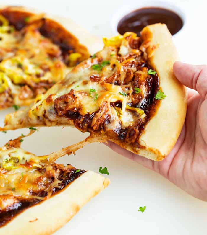 A hand pulling up a warm slice of BBQ Chicken Pizza with a cup of BBQ Sauce in the background.