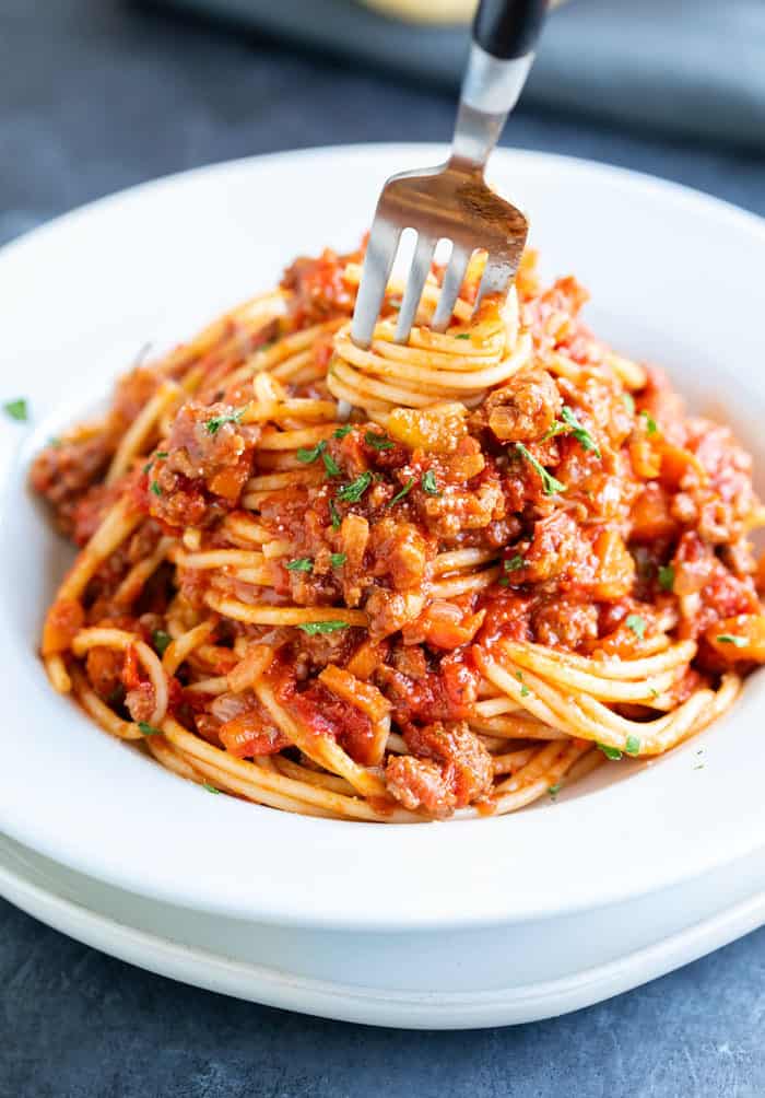 A white bowl filled with spaghetti bolognese with a fork at the top twisting the spaghetti.