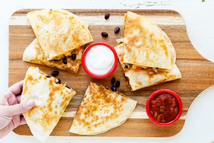 Wooden cutting board with crispy chicken quesadillas with sour cream and salsa. A hand is reaching for a quesadilla.