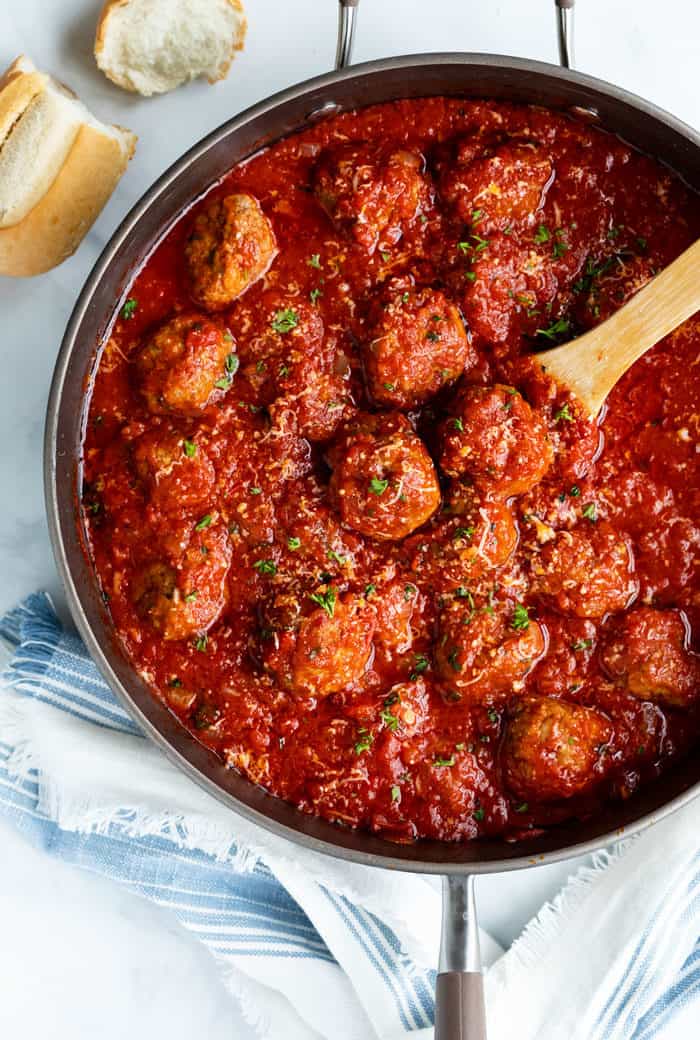 A large skillet filled with marinara sauce and meatballs with a wooden spoon on a white marble surface and a white and blue kitchen towel.