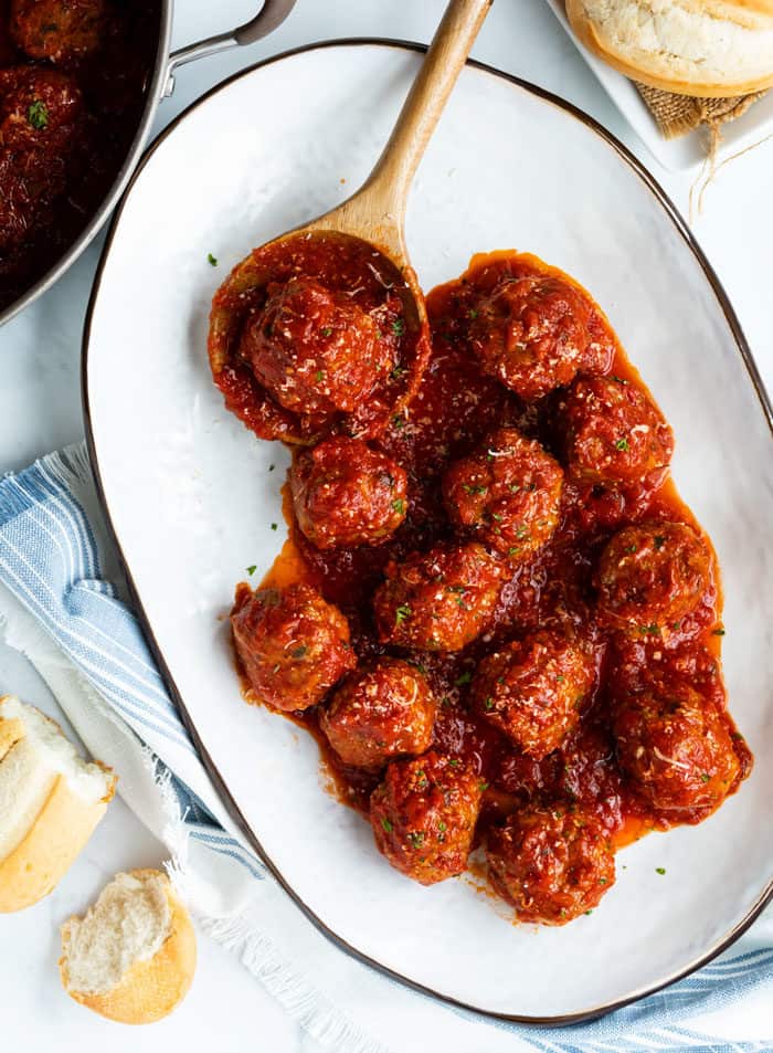 A white serving platted with a wooden spoon, marinara sauce, and meatballs on a white surface with bread around it.