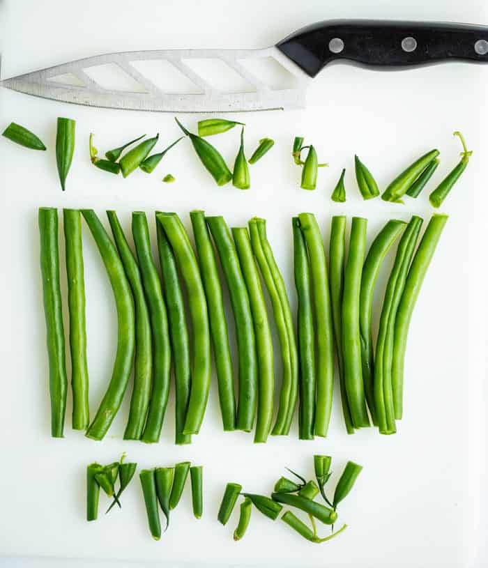 A line of fresh green beans on a white surface with a knife and the stems cut off on each side.
