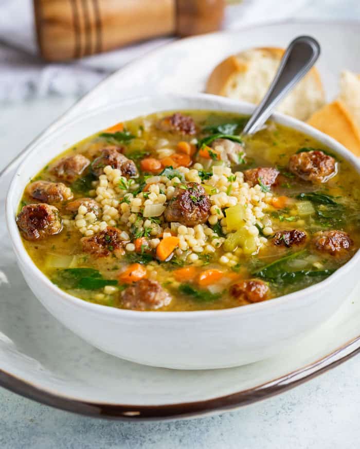 A white bowl of Italian wedding soup on a white tray with a spoon in it and rolls in the background.