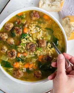 A white bowl full of Italian Wedding Soup with a hand holding a spoon.