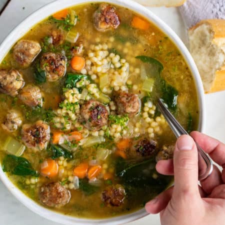 A white bowl full of Italian Wedding Soup with a hand holding a spoon.