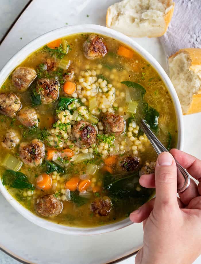 Overhead view of a white bowl of Italian wedding soup with a hand holding a spoon in it and rolls next to the bowl.