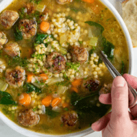 Overhead view of a white bowl of Italian Wedding Soup with a hand holding a spoon.