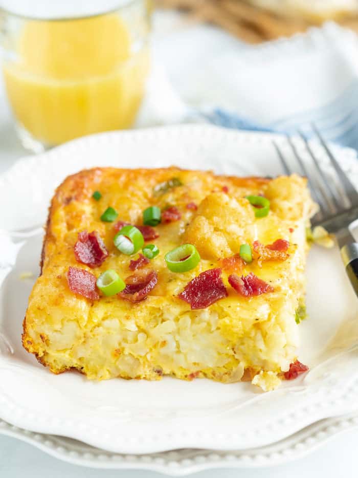 A slice of tater tot breakfast casserole on a white plate with a fork and orange juice in the background.