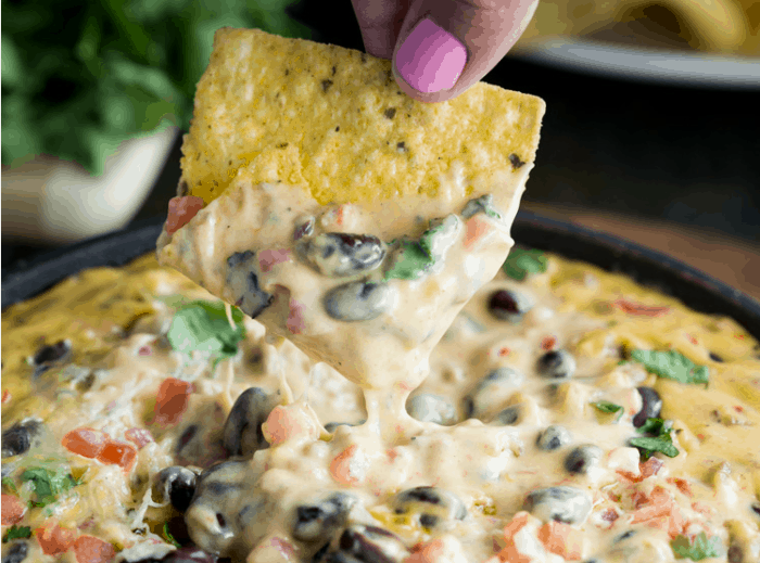 A hand dipping a tortilla chip into a cast iron full of loaded cowboy queso dip.
