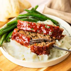 A white plate with Cracker Barrel Meatloaf on top of mashed potatoes with a fork. Green beans in the background.