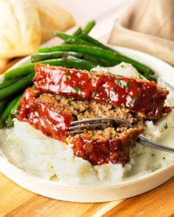 A white plate with Cracker Barrel Meatloaf on top of mashed potatoes with a fork. Green beans in the background.