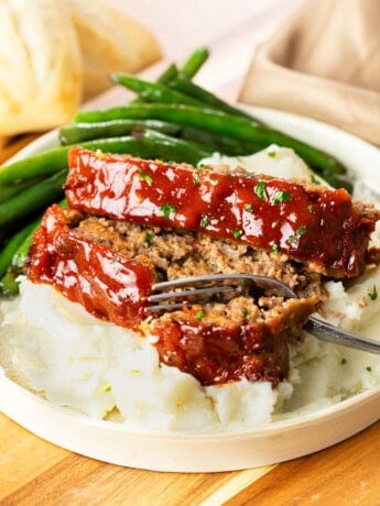 A white plate with Cracker Barrel Meatloaf on top of mashed potatoes with a fork. Green beans in the background.