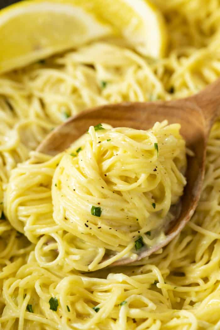 A wooden spoon holding a scoop of creamy lemon pasta in a pot with lemons in the background.