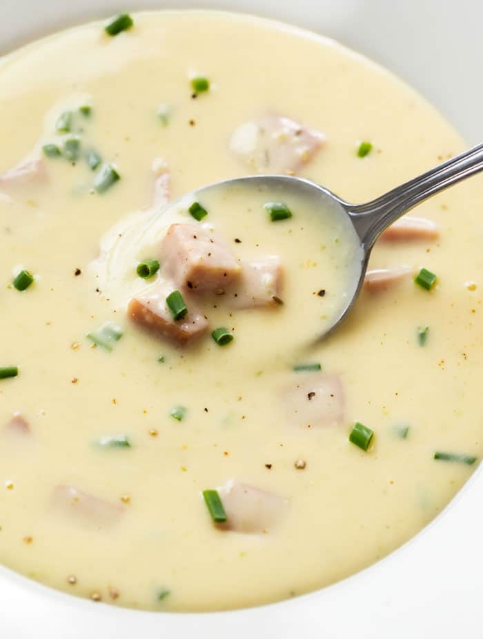 A spoon scooping up ham and potato soup with chives from a white bowl.