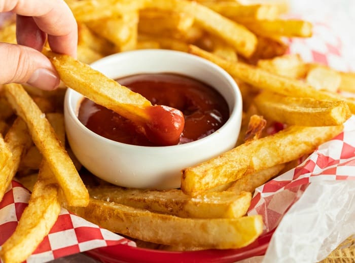 A hand dipping golden french fries into a white ramekin filled with ketchup.
