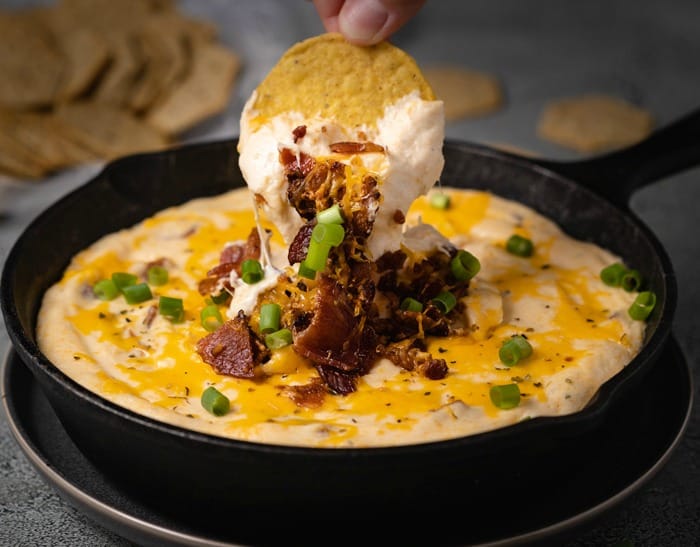 A hand dipping a tortilla chip into a cast iron skillet filled with warm bacon ranch cheese dip.