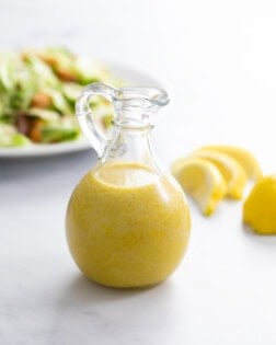 A glass bottle filled with homemade Caesar Dressing with a salad and lemon slices in the background.