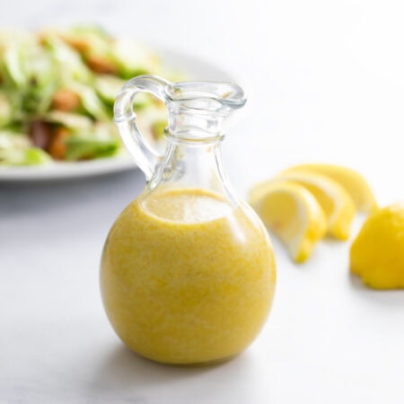 A glass bottle filled with homemade Caesar Dressing with a salad and lemon slices in the background.