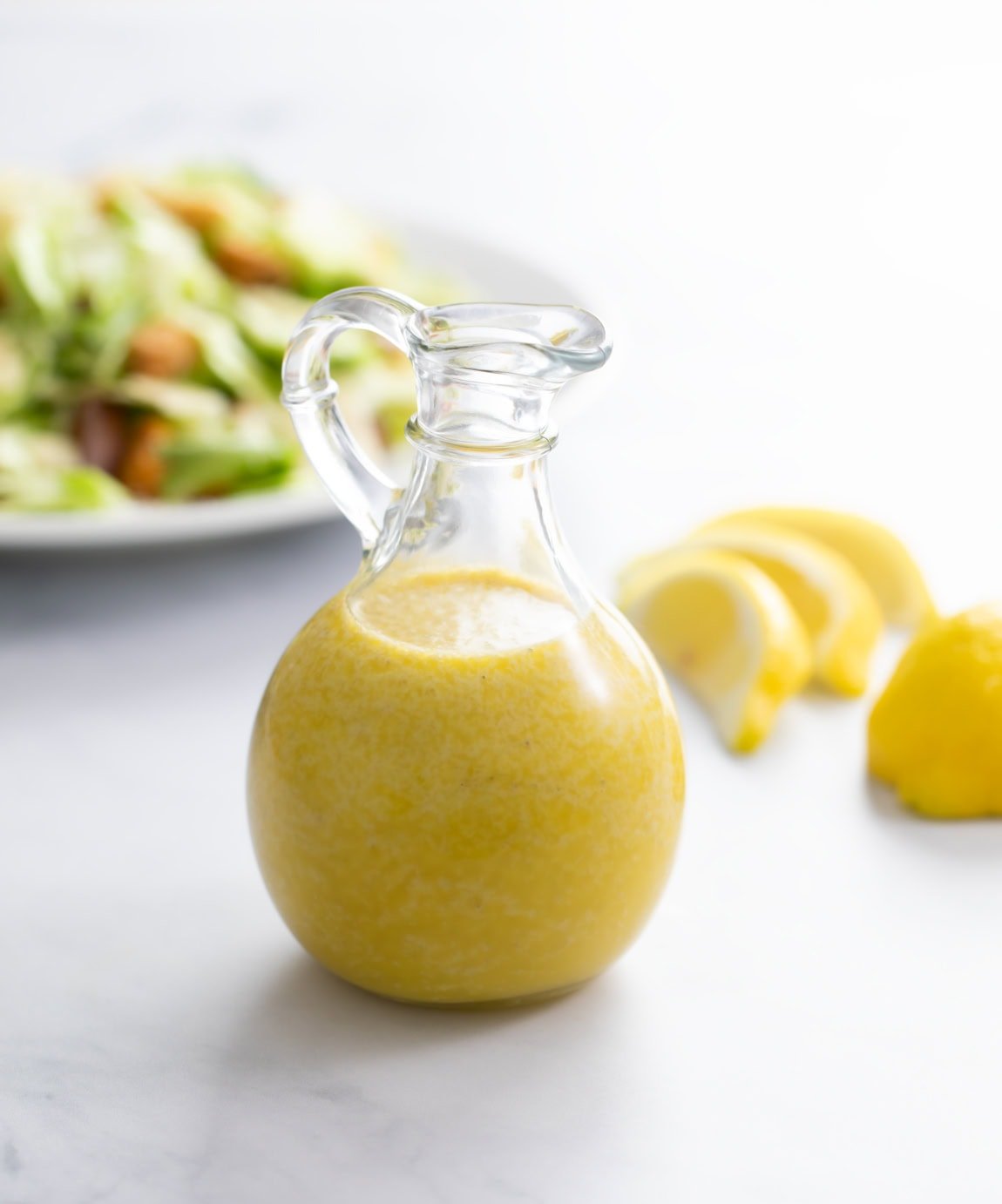 A glass bottle filled with homemade Caesar Dressing with a salad and lemon slices in the background.