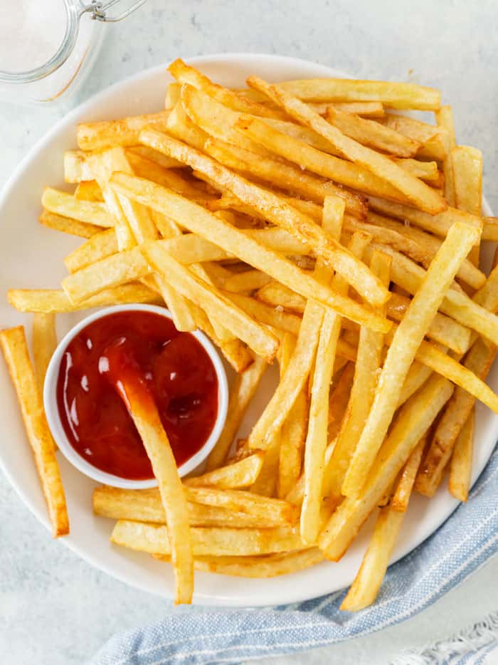 A plate of golden french fries on a white plate with a bowl of ketchup for dipping.