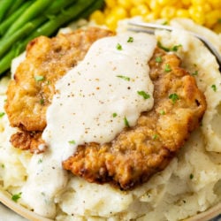 Crispy Chicken Fried Steak on a bed of mashed potatoes with white gravy on top and vegetables in the back.