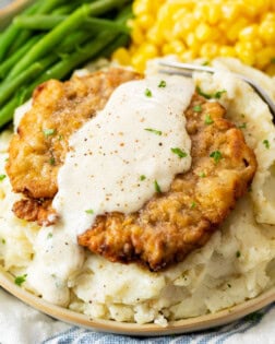 Crispy Chicken Fried Steak on a bed of mashed potatoes with white gravy on top and vegetables in the back.