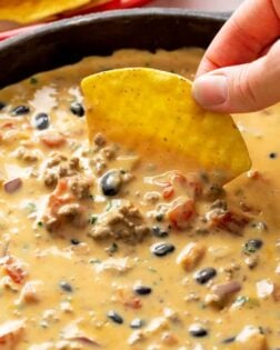 A hand dipping a tortilla chip into a skillet filled with Queso Dip with black beans, beef, and tomatoes.