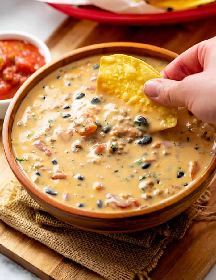 A bowl filled with queso dip with black beans and tomatoes, with a hand dipping a tortilla chip in it.