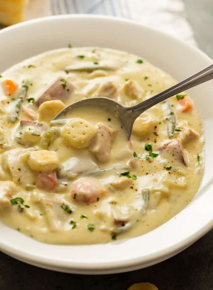 A white bowl of creamy chicken stew topped with oyster crackers and a spoon scooping it up.