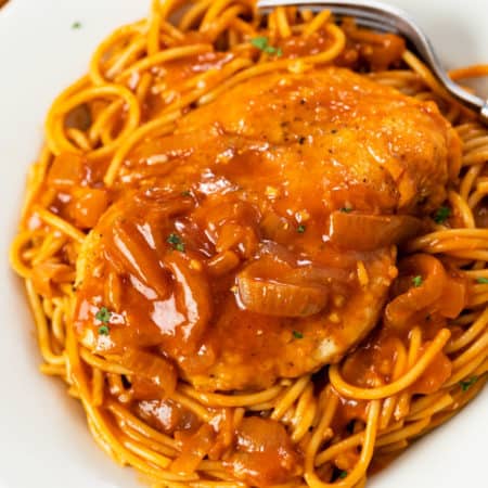 Chicken breast on top of a pile of spaghetti with a red chicken scallopini sauce with sliced onions and parsley.
