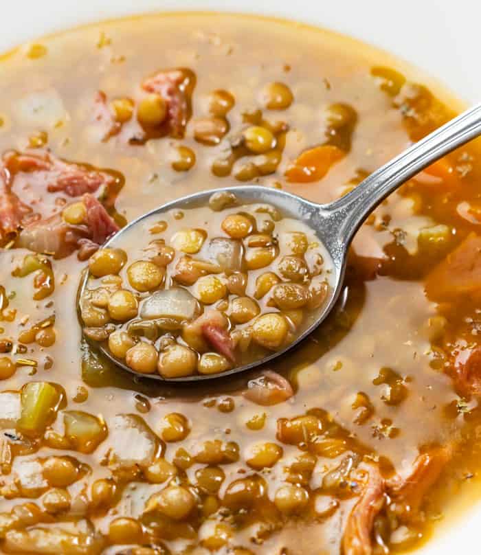A metal spoon full of cooked lentils in a bowl of lentil soup with ham.