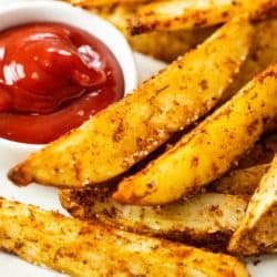 A pile of crispy oven baked potato wedges next to a white bowl of ketchup.