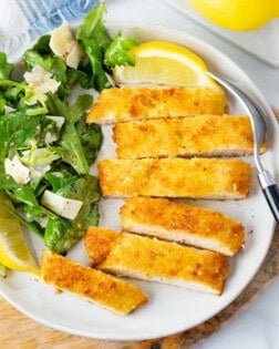 Chicken Milanese cut into slices on a plate with lemon wedges, fresh greens, and Parmesan shavings.