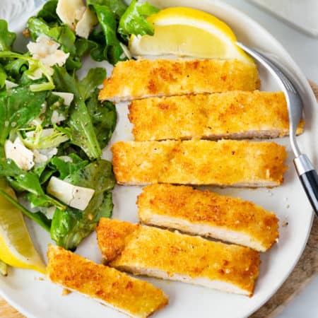 Chicken Milanese cut into slices on a plate with lemon wedges, fresh greens, and Parmesan shavings.