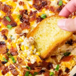 A hand holding a crusty piece of bread and dipping it into hot corn dip topped with bacon.