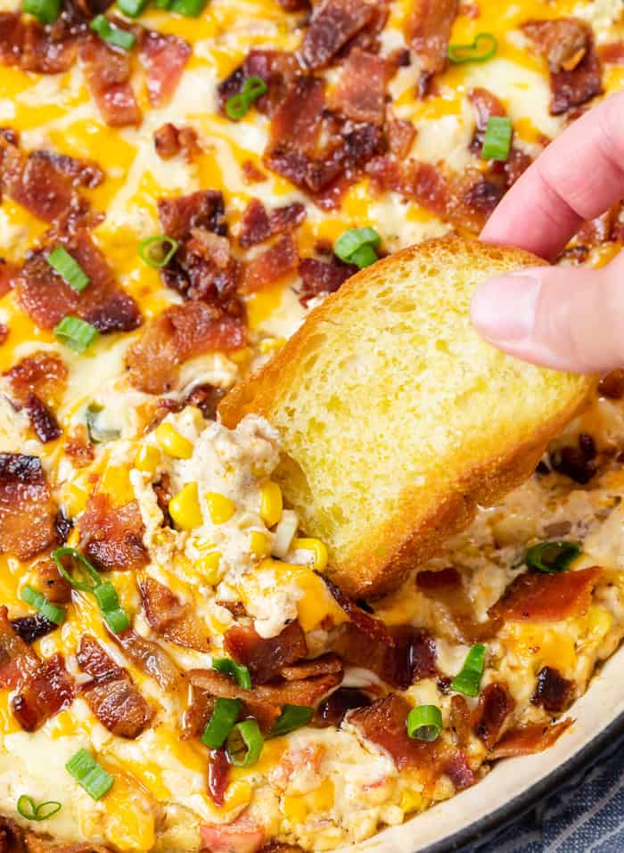 A hand holding a crusty piece of bread and dipping it into hot corn dip topped with bacon.